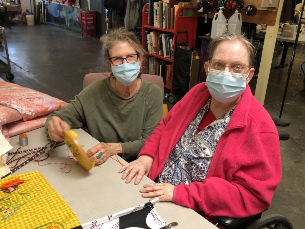 Volunteer at NIAD. NIAD Volunteer seated next to a NIAD artist in the studio. They pause work on their art project to look up at the camera.