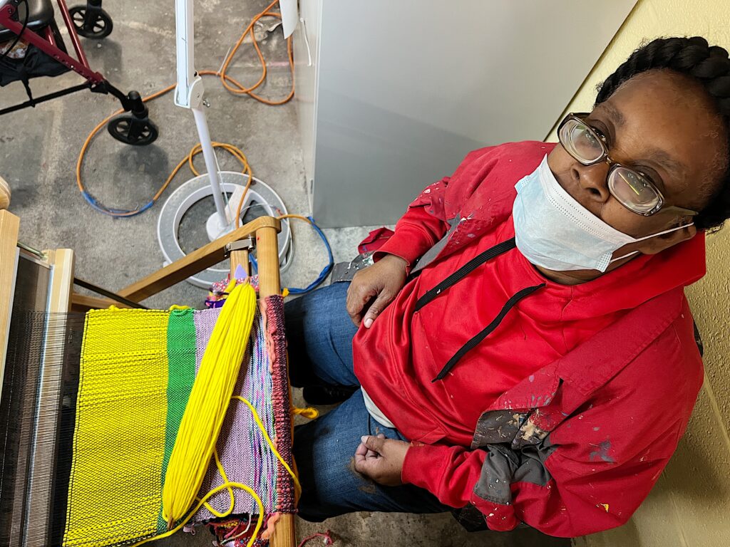 a woman with black braids and a mask on, wearing a red jacket, sits in front of a loom working with yellow, lavender and green yarn. she is looking up at the camera. There are many ways to give to NIAD Art Center.
